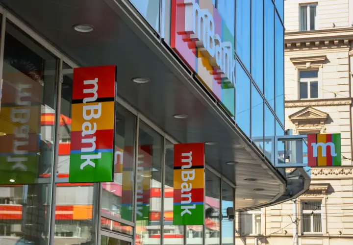 Baner mBank z kolorowym logo i napisem "mBank" na tle nowoczesnego budynku.