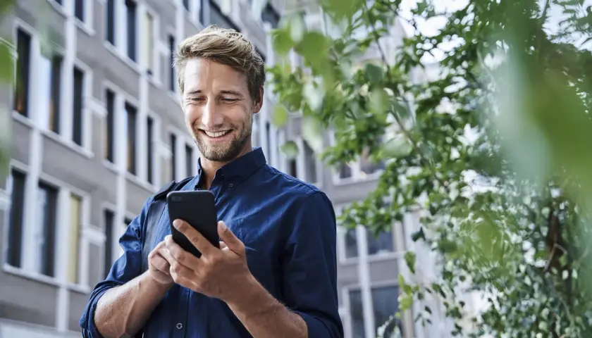 Uśmiechnięty mężczyzna w granatowej koszuli korzysta z telefonu na tle budynku i zieleni.
